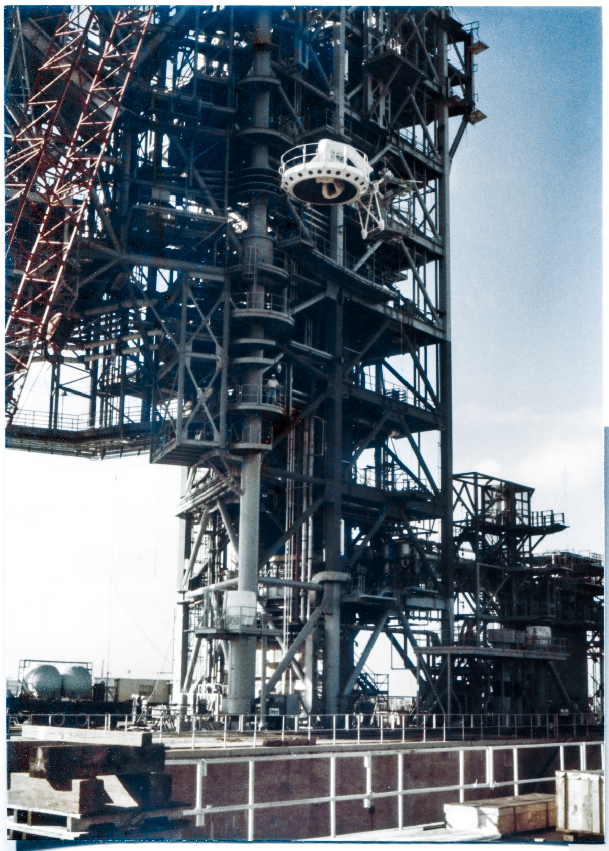 Image 119. At Space Shuttle Launch Complex 39-B, Kennedy Space Center, Florida, the white flying-saucer-looking ”Beanie Cap” of the External Tank Gaseous Oxygen Vent System Tip Assembly can be seen rising skyward with the Fixed Service Structure directly behind it, being lifted by the crane to its final elevation nearly 250 feet above the Pad Deck, where it will be attached by Ivey Steel's Union Ironworkers to the end of the GOX Arm, which is attached to the FSS out of frame above, and not visible in this photograph. Photo by James MacLaren.